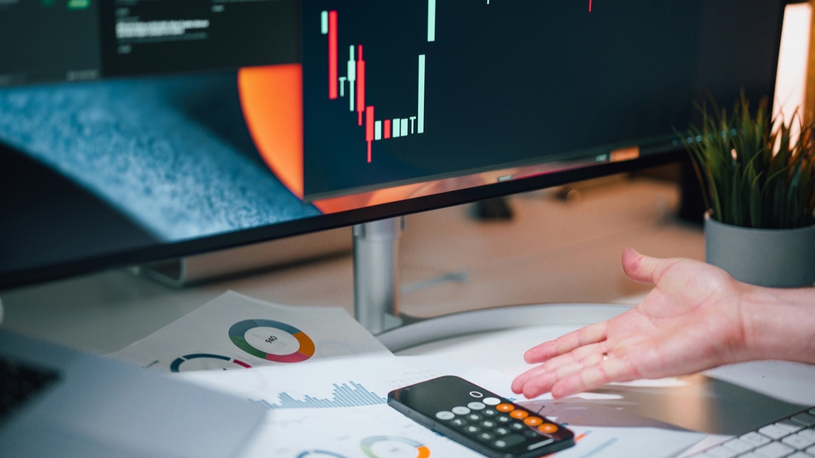 Mão aponta para uma calculadora sobre relatórios financeiros em frente a um monitor com gráfico