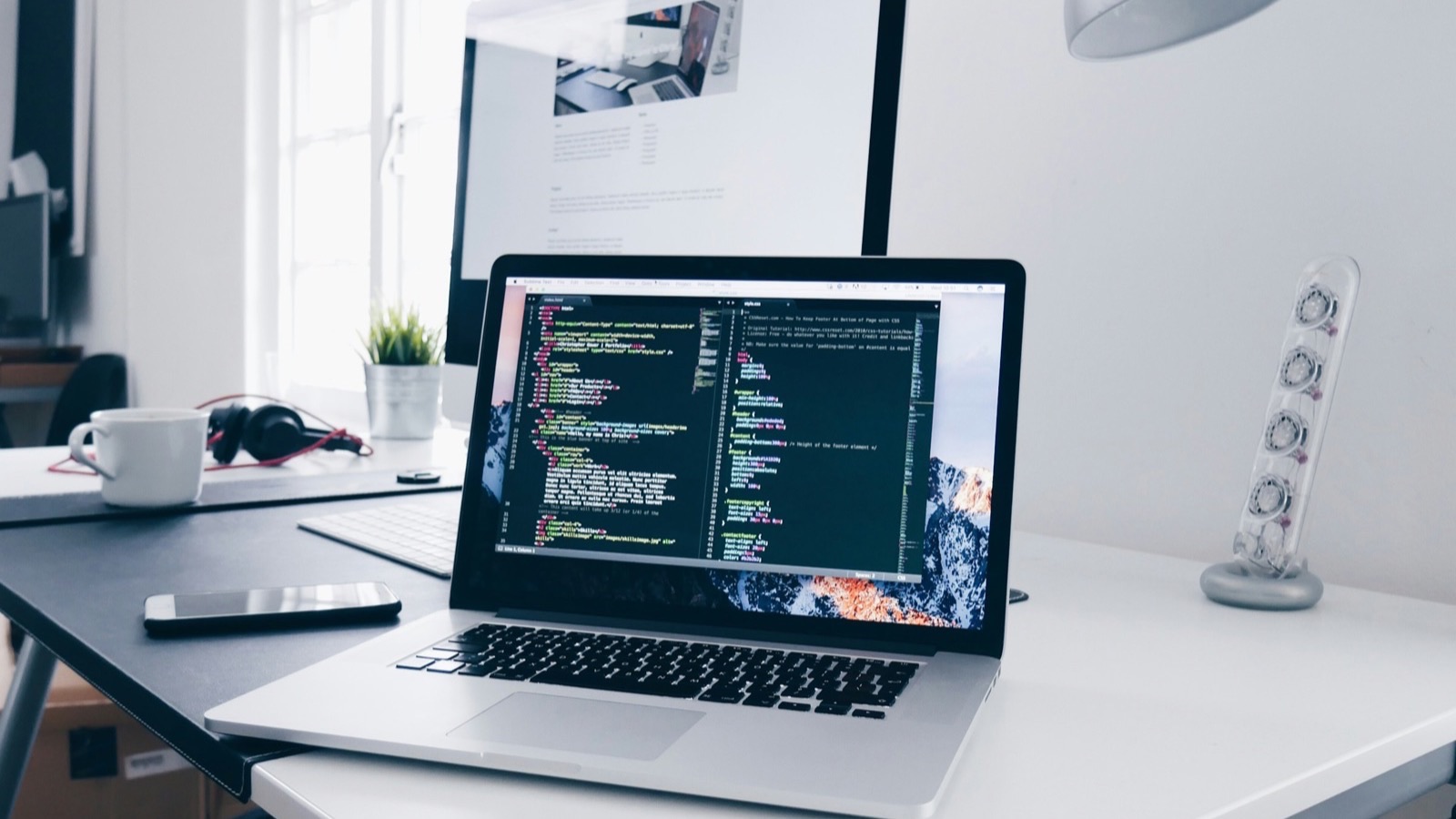 Notebook com código de programação aberto na tela, sobre uma mesa de escritório com monitor ao fundo