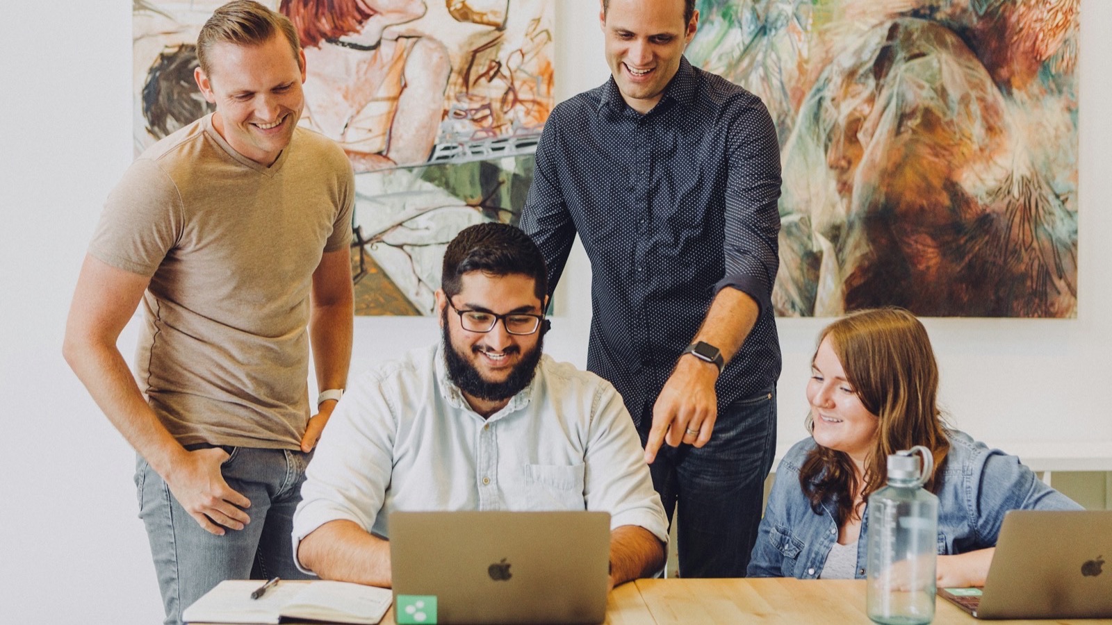 Equipe de quatro pessoas sorrindo enquanto analisa um projeto em um notebook sobre uma mesa de escritório