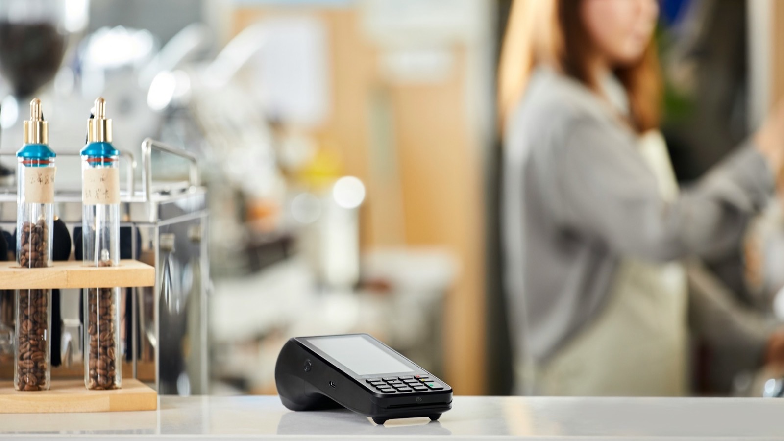 Maquininha de cartão sobre o balcão de uma cafeteria, com atendente ao fundo