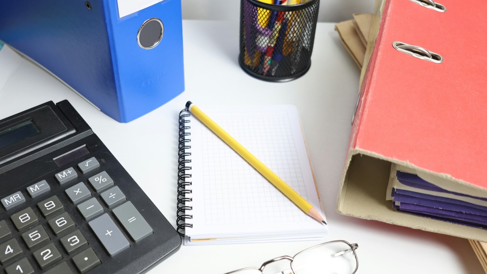 Calculadora, caderno quadriculado com lápis, pastas de arquivo e óculos sobre uma mesa de escritório